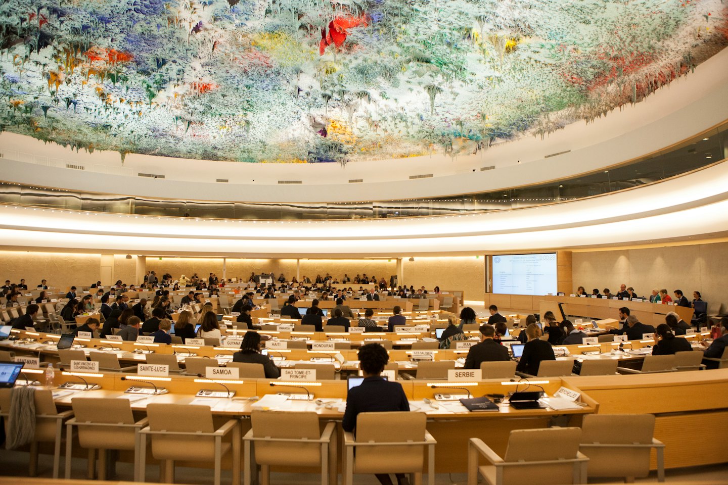 Delegati in una moderna sala conferenze partecipano a una riunione sotto un soffitto a cupola colorato.