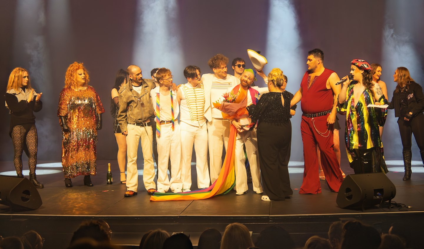 Artisti festeggiano su un palco teatrale illuminato con una bandiera arcobaleno.